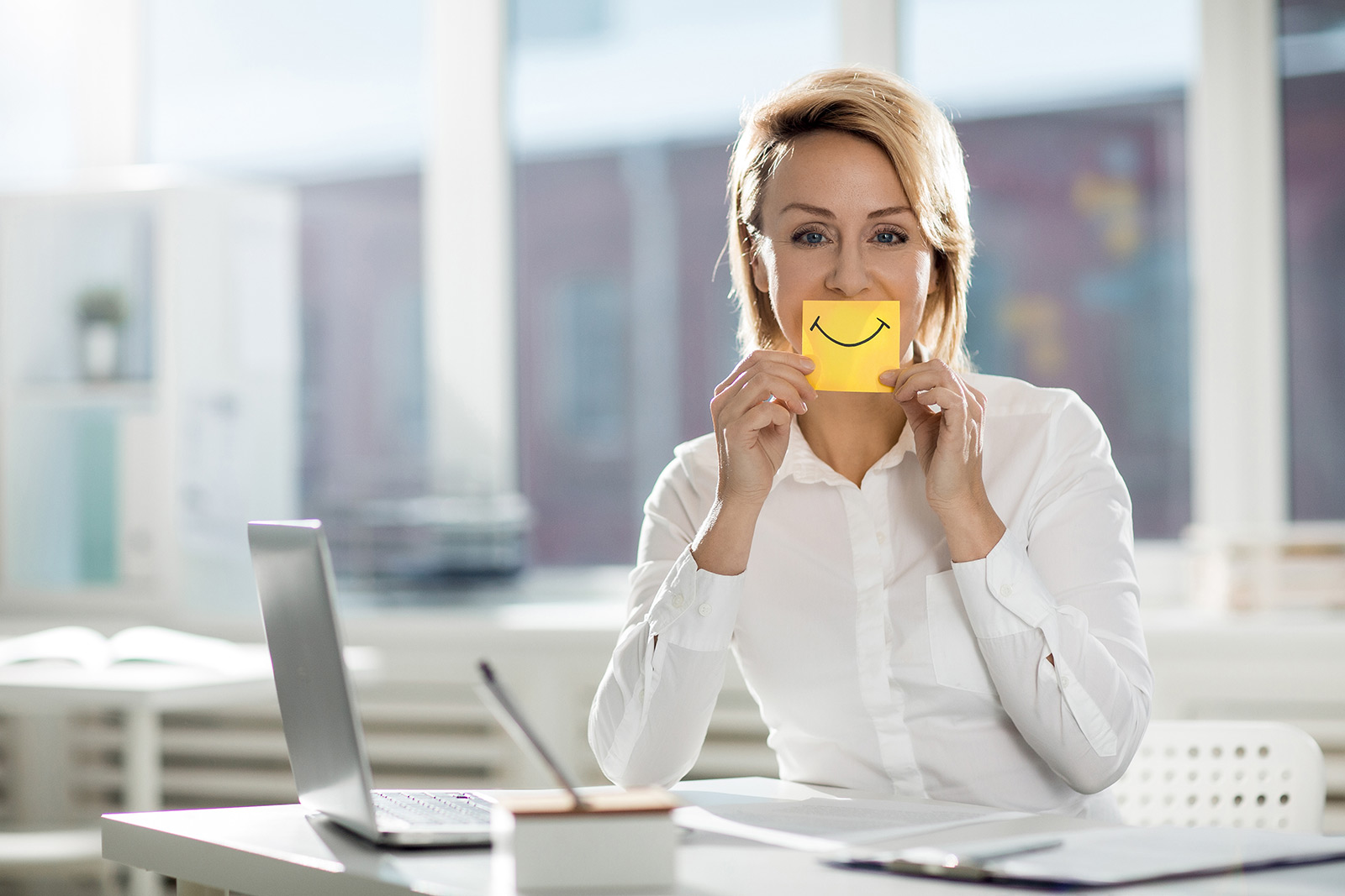 A home care agency owner at her laptop. Financial Health of her business is good  and so is Client Satisfaction, so she is holding up a &lsquo;smiley&rsquo; mouth drawn on a post-it held over her own mouth.